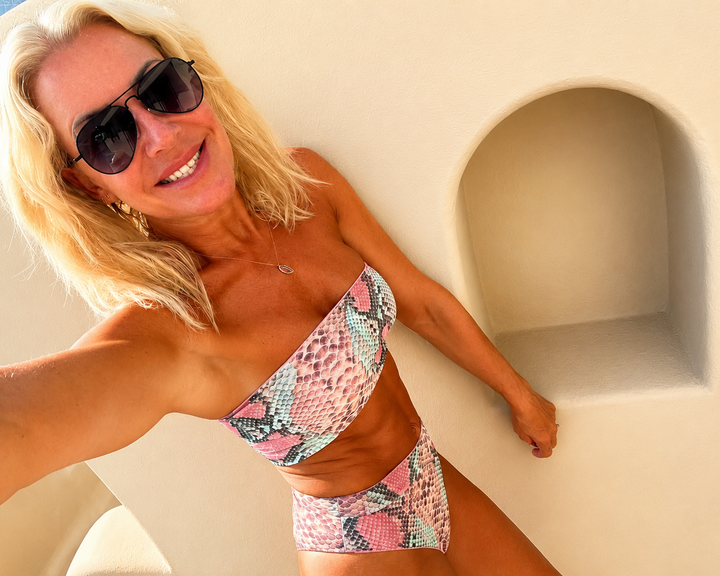 A blonde woman wearing a pink, blue, and white snakeskin-patterned bikini top and matching high-waisted bottoms, posing in a selfie on a sunny day.